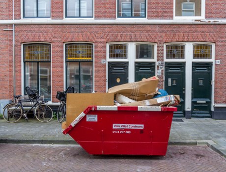 A red skip bin with a white and red safety sticker, positioned on the pavement in front of a row of red brick terraced houses with white window frames and dark green front doors, is filled with various household waste materials including cardboard boxes, paper, and plastic packing, some of which are partially protruding from the top. The skip is situated on a residential street in Cheshunt, with a few bicycles parked against the wall to the left side of the image. The scene appears outdoors during daylight with overcast weather, and the overall setting suggests it is part of a house clearance or rubbish removal service managed by House Clearance Cheshunt, as indicated by local environmental standards and waste disposal practices typical of the area.