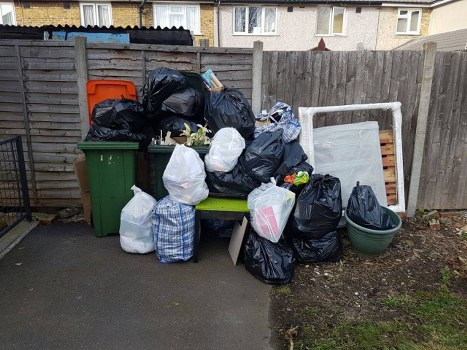 A photograph depicting a collection of domestic rubbish piled against a weathered wooden fence in an outdoor setting. The waste includes black bin bags of varying sizes, some tightly knotted and others loosely filled, placed on and around a green plastic bench. In addition, there are white and blue checkered plastic bags, a large green waste bin, and a few loose items such as a small cardboard box and a plastic container. The rubbish appears to consist of household waste materials, primarily bags filled with miscellaneous debris, positioned on a paved surface adjacent to soil with patches of grass. The background features a wooden fence with horizontal slats, behind which parts of residential buildings are visible, suggesting this is a property in a suburban area consistent with the vicinity of Cheshunt, Hertfordshire. The overall scene is neutral in tone, typical of a temporary rubbish disposal area, and aligns with services offered by House Clearance Cheshunt for rubbish removal and clearance in the local community.