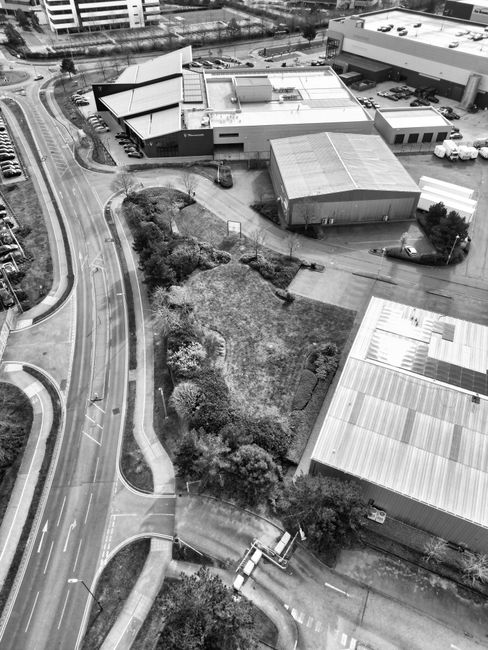 An aerial black-and-white photograph showing a complex of industrial and commercial buildings situated near curving roads. In the foreground, there is a landscaped area with grass, trees, and bushes,